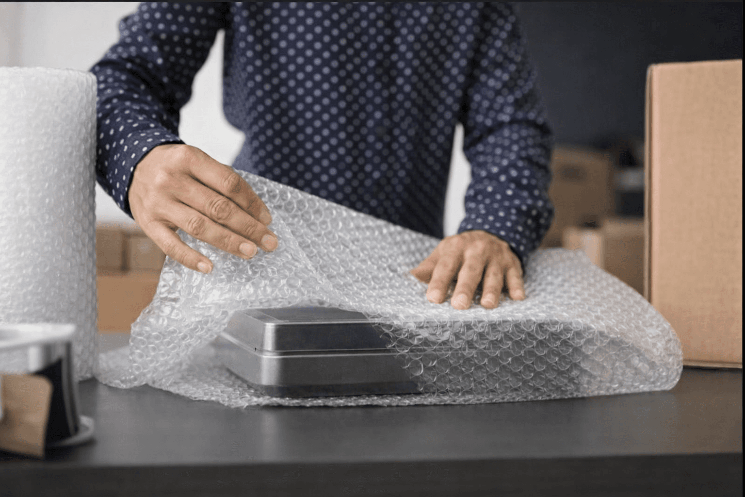A person holding a roll of small bubble wrap in a warehouse in Blackburn, packing a fragile ceramic mug into a cardboard box.